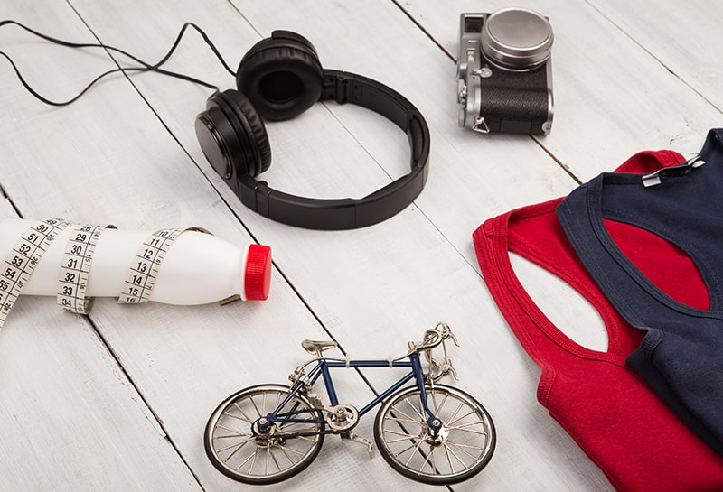 een afbeelding van een rode en zwarte fietsvest, een speelgoedfiets, een witte plastic waterfles omwikkeld met meetlint, een koptelefoon en een vintage camera op een houten tafel